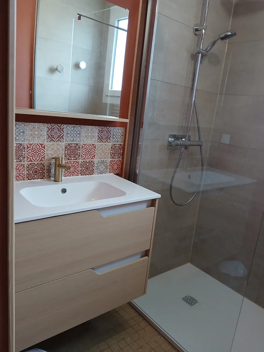 Une salle de bains refaite : douche vitrée, meuble vasque en bois clair, carreaux à motifs rouges